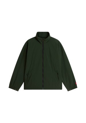 Giovane uomo con giacca e pantaloni verde scuro, camicia viola e cappellino verde con logo rosso, in piedi su sfondo bianco uniforme.