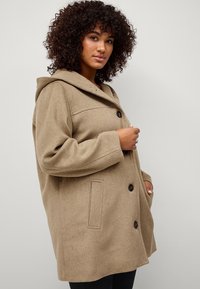Beige wollen blend jas met capuchon, met een dubbelgevoerde ontwerp, grote knopen en zijzakken. Zachte textuur, iets oversized pasvorm.