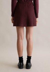 Burgundowa wełniana spódnica mini z prostym dołem i tylnym zamkiem błyskawicznym. Modelka nosi czarne skarpetki i masywne czarne buty, nogi widoczne.