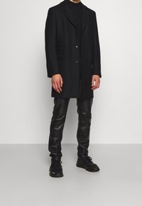 Homme portant un pardessus noir, un col roulé noir, un pantalon en cuir noir et des baskets noires, debout devant un fond blanc uni.