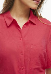 Košeľa s krátkym rukávom a golierom v živej ružovej farbe. Vyrobená z textúrovanej látky, s vreckom a šiestimi gombíkmi na zapínanie.