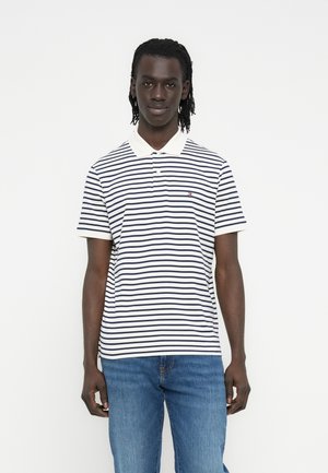 Fonott hajú férfi, aki fehér és tengerészkék csíkos galléros pólót és kék farmert visel, fehér háttér előtt áll.