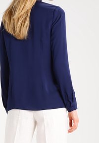 Blouse bleu marine en tissu lisse, avec des manches longues et des poignets à boutons, et une coupe droite à l'ourlet. Pantalon blanc visible en dessous.