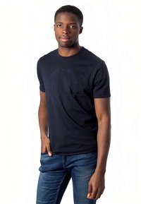 Νεαρός άνδρας ντυμένος με σκούρο ναυτικό μπλε μπλουζάκι Armani Exchange και μπλε τζιν, στέκεται με το ένα χέρι στην τσέπη, μπροστά σε λευκό φόντο.