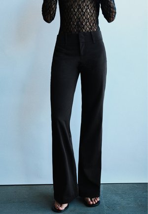 Personne portant un pantalon noir évasé, un haut en dentelle noire transparente et des sandales noires à bouts ouverts, debout devant un fond clair uni.