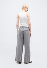 Άτομο με κοντά ξανθά μαλλιά, ντυμένο με λευκό αμάνικο τοπ, χαλαρό γκρι παντελόνι και μαύρες μυτερές γόβες, στέκεται μπροστά σε ένα απλό φόντο.