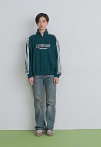 Mlad človek stoji ob enostavni steni, oblečen v temno zeleno Adidas jopico, modre kavbojke in rjave čevlje.