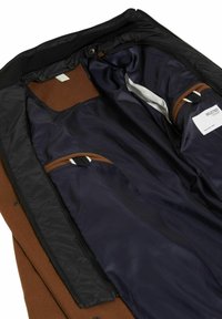 Brun frakk med svart fôr, med en glatt marineblå innside. Inkluderer to innerlommer med kontrastdetaljer og et merke.