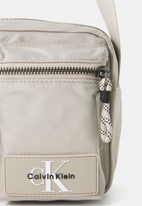 Compact beige crossbody bag made from smooth nylon. Features a front zip pocket, textured pull tab, and a logo plate with raised lettering.