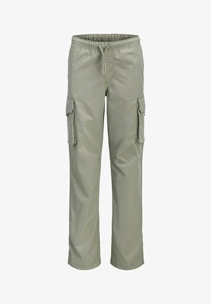 Adolescent aux cheveux bouclés portant une chemise bleu marine à manches courtes sur un haut blanc à manches longues, un pantalon cargo clair, et des baskets, posé sur un fond uni.