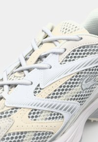 Gros plan sur une chaussure de sport en mesh blanc et beige avec des lacets blancs et des surpiqûres synthétiques texturées.