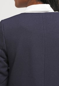 Mörkblå texturerad jacka med skräddarsydd passform, med en vit krage. Tyget har ett vävt mönster och synliga sömdetaljer.