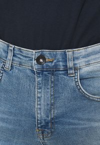 Lyseblå denimjeans med høj talje, præget af en sort knaplukning, gule syninger og forlommer. Teksturen fremstår let slidte.