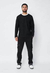Sweatshirt noir à manches longues avec un col rond blanc et un pantalon cargo noir avec poches et un look usé. Accents blancs sur le pantalon.