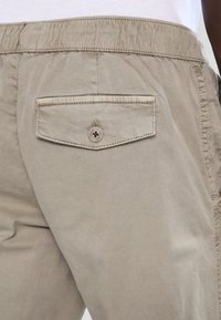 Beige bomullsshorts med elastisk midjeband och en bakficka med knappstängning. Tyget har en jämn, matt yta.