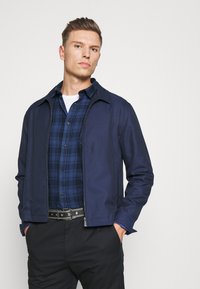 Mornarska zip-up jakna z gladko teksturo, s konico ovratnikom; prekrita z modrim karirastim srajcem, dopolnjena s črnimi hlačami in pasom s študami.