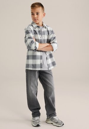 Grijze en witte geblokte flanellen shirt, dichtgeknoopt, gecombineerd met grijze spijkerbroek en sportsneakers. Achtergrond is eenvoudig, waardoor de outfit benadrukt wordt.