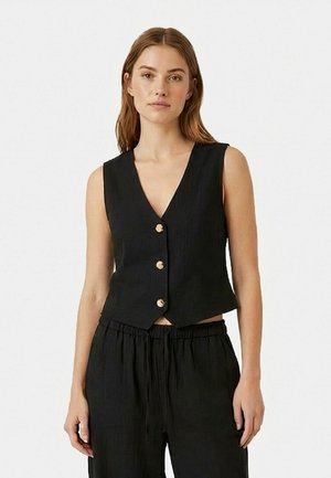 Nő fekete, ujjatlan, gombos mellényben, V-nyakú, hozzáillő laza fekete nadrágban, fehér egyszínű háttér előtt áll.