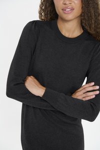 Görög hajú nő, fekete, hosszú ujjú kötött pulóverruhában, puffos vállrésszel, karját összefonta.