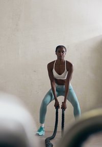 Kvinde i sportstøj udfører battle rope øvelse i et minimalistisk, lyst træningslokale med betonvægge.