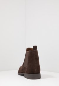 Botín Chelsea de ante marrón con paneles elásticos en los laterales y una lengüeta en el talón. Cuenta con una suela de goma lisa y un diseño hasta el tobillo.