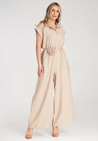 Beżowy kombinezon z krótkim rękawem, z kołnierzykiem, przewiązany w talii i zdobiony materiałowym kwiatem; szerokie nogawki w miękkim, teksturowanym materiale.