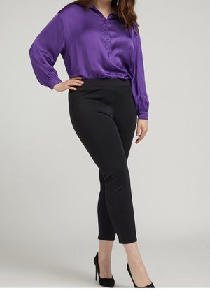 Femme portant un chemisier en satin violet, un pantalon noir ajusté aux chevilles et des chaussures à talons noirs, debout avec une main sur la hanche.