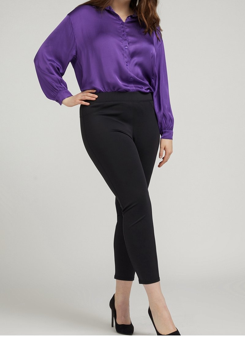 Femme portant un chemisier en satin violet, un pantalon noir ajusté aux chevilles et des chaussures à talons noirs, debout avec une main sur la hanche.