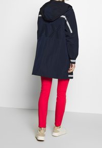 Marine windbreaker met capuchon, heeft een groot "AX" logo op de achterkant, reflecterende strepen op de mouwen, gecombineerd met felroze broeken en beige sneakers.