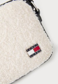 Väike valge kiharate fliisist kott musta äärisega ja metallse Tommy Jeans logo, millel on punane, valge ja tumesinine ristkülik.