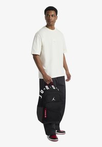 Rucsac Jordan negru cu logo alb și accente roșii, purtat de o persoană îmbrăcată într-o cămașă crem și pantaloni negri, evidențiat pe un fundal alb.