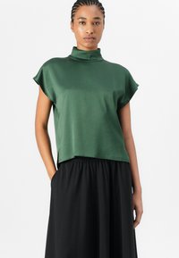 Haut vert à col montant en tissu lisse avec des manches courtes, associé à une jupe noire avec une taille élastique et des poches.