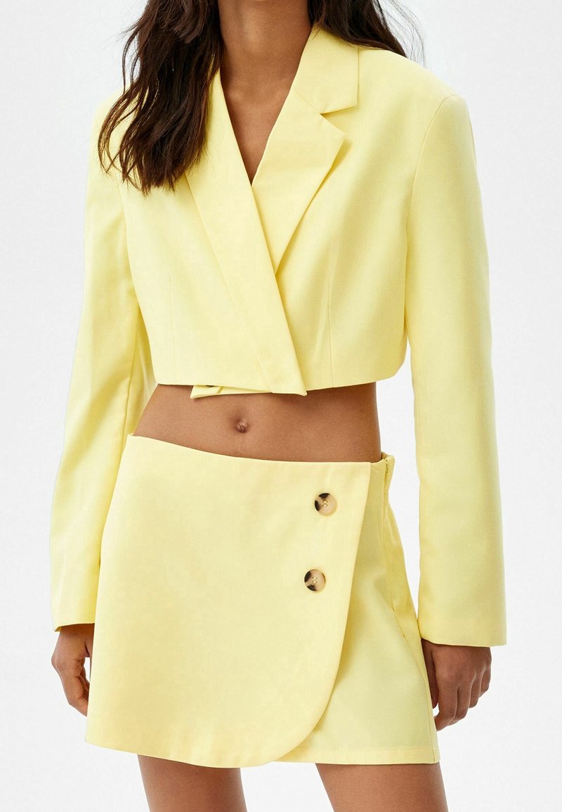 Woman wearing a pale yellow cropped blazer with long sleeves and a matching mini skirt with two buttons on one side.