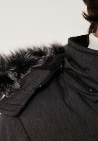 Primer plano de un abrigo de invierno gris oscuro con un cuello alto y ribete de piel sintética negra alrededor de la zona de los hombros, usado sobre un jersey de cuello alto blanco.