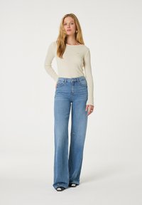 Beige geribbelde gebreide top met lange mouwen, gecombineerd met lichtblauwe wijde jeans met kleurrijke geborduurde accenten. Effen achtergrond; geen aanvullende details.