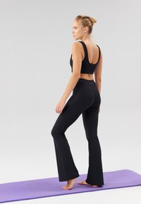Top sport crop negru cu bretele late, asortat cu pantaloni negri evazați pentru yoga. Ambele articole sunt realizate dintr-un material neted și elastic, prezentate pe un covoraș mov.