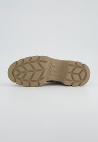 Texturierte beige Schuhsohle mit tiefen Profilmustern für besseren Halt; verfügt über Branding im Bereich des Fußgewölbes und eine glatte Oberfläche.