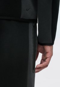 Czarna i szara sportowa kurtka o gładkiej strukturze, z ukrytą kieszenią na zamek oraz subtelnym logo Nike na górnej części.