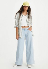 Camicia a righe con bottoni, top corto bianco, jeans a gamba larga di un lavaggio chiaro e cappellino giallo con grafiche di cactus. Outfit casual con una vestibilità comoda.