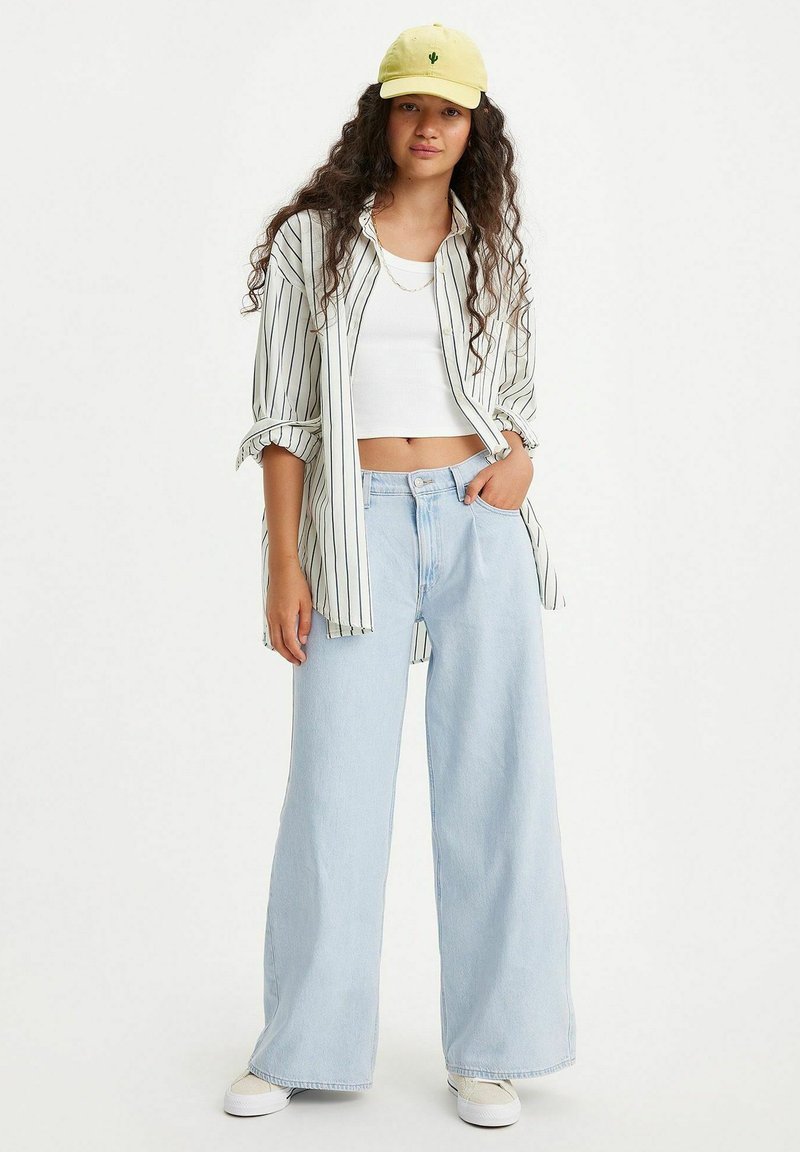 Camicia a righe con bottoni, top corto bianco, jeans a gamba larga di un lavaggio chiaro e cappellino giallo con grafiche di cactus. Outfit casual con una vestibilità comoda.