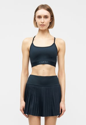 Femeie purtând un top sport negru scurt cu bretele subțiri și o fustă plisată neagră cu talie înaltă, stând în fața unui fundal simplu, deschis la culoare.