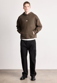 Rudas gobtuvu megztinis su priekiu kišene ir logotipu, derinamas su juodomis kargo kelnėmis su šoninėmis kišenėmis ir laisvu kirpimu.