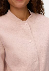 Chaqueta de tela fieltro en rosa claro, con cuello redondo, que cuenta con cuatro botones a presión rosas y una superficie texturizada.