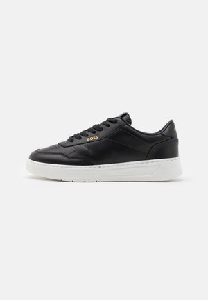 Zapatilla deportiva de cuero negro con suela de goma blanca, que presenta un sutil logo dorado en el lateral y un diseño clásico con cordones.