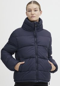 Námořnická puffer bunda s vysokým límcem, zipem vpředu a bočními kapsami. Má prošívaný design, zaoblený lem a elastické manžety.