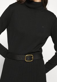 Pull à col roulé noir en matériau doux, avec des manches longues et un logo discret. Associé à une ceinture noire texturée avec boucle dorée.