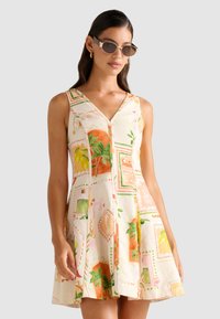 Modellen har på seg en ermeløs, mønstret kjole med blomster- og fruktmotiver. Hun poserer med store solbriller foran en lys bakgrunn.