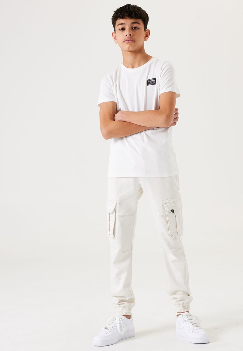 Witte t-shirt met ronde hals en zwart logo-embleem, gecombineerd met lichtbeige cargobroek met zijzakken en elastische cuffs, en witte sneakers.