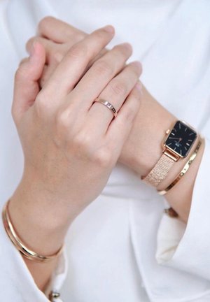 Deux mains serrées, l'une portant une montre en or rose, un bracelet assorti et une bague en or rose, sur un fond de chemise blanche.