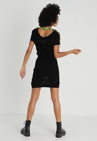 Robe noire à manches courtes, avec un dos échancré et une jupe texturée. Accentuée par une lanière jaune vif le long des épaules.